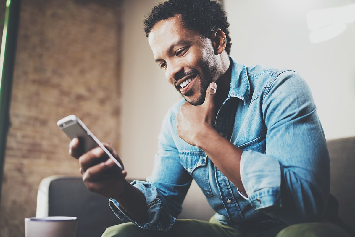 Happy bearded African-American businessman using phone while sitting on sofa at his modern home