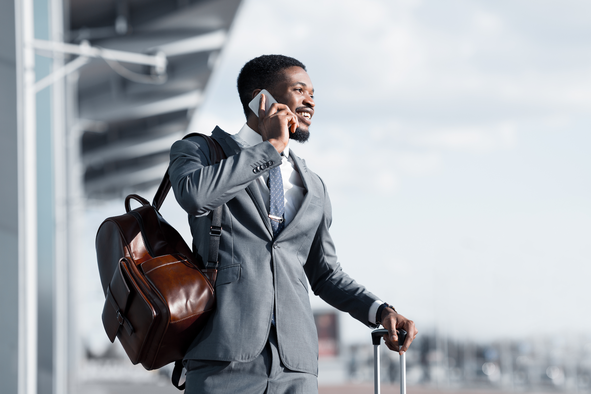 A Black man wearing a tailored suit talks on a phone at an airport while waiting for a taxi. He has a leather backpack slung over one shoulder and the handle for a rolling suitcase in the other.