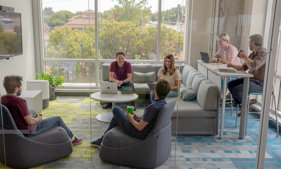 ​ Six Brinqa team members work in a shared lounge area, talking and smiling.