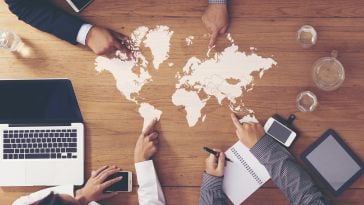 A photo of a desk with a map of the world with four employees pointing at various locations.