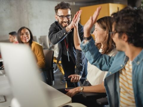 Team members seated in front of computer monitors and high-fiving each other in office